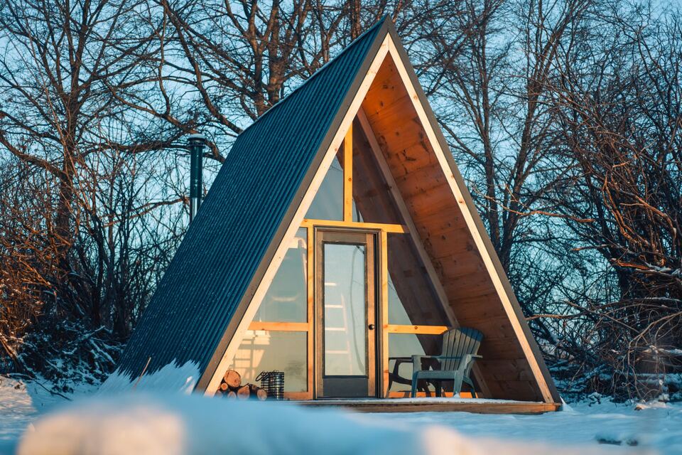 Finished A-frame cabin with a glazed front and dark metal roof in a snowy wooded setting