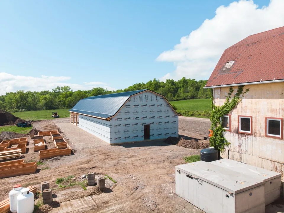 Pole barn under construction with house wrap and a metal roof beside an existing barn